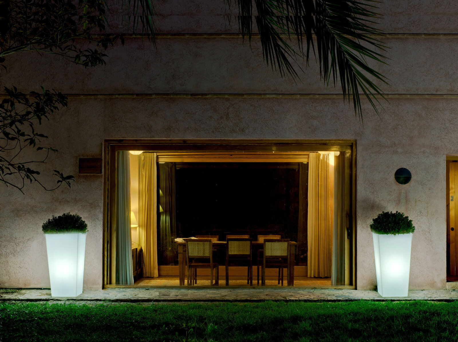 Zwei beleuchtete Blumentöpfe Melisa 50 vor einem großen Fenster, nachts im Garten platziert, mit Blick auf das Haus.