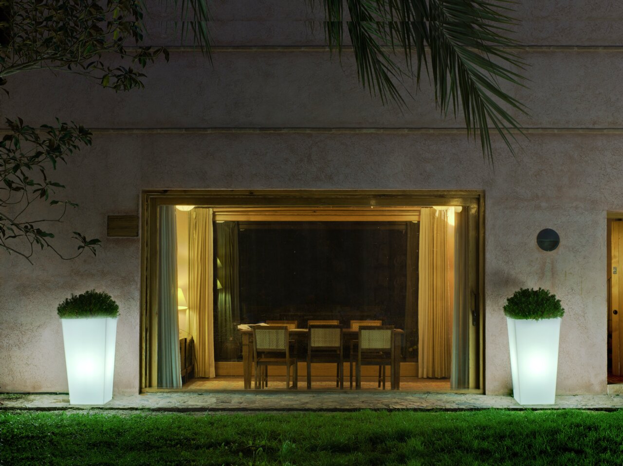Zwei beleuchtete Blumentöpfe Melisa 50 vor einem großen Fenster, nachts im Garten platziert, mit Blick auf das Haus.