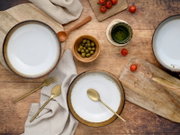 Rustikales Tisch-Setting mit drei weißen Keramikschalen mit braunem Rand, goldfarbenem Besteck, Holzlöffel, Schale mit grünen Oliven, Cherrytomaten und grünem Glas auf Holzuntergrund, Draufsicht (von oben)