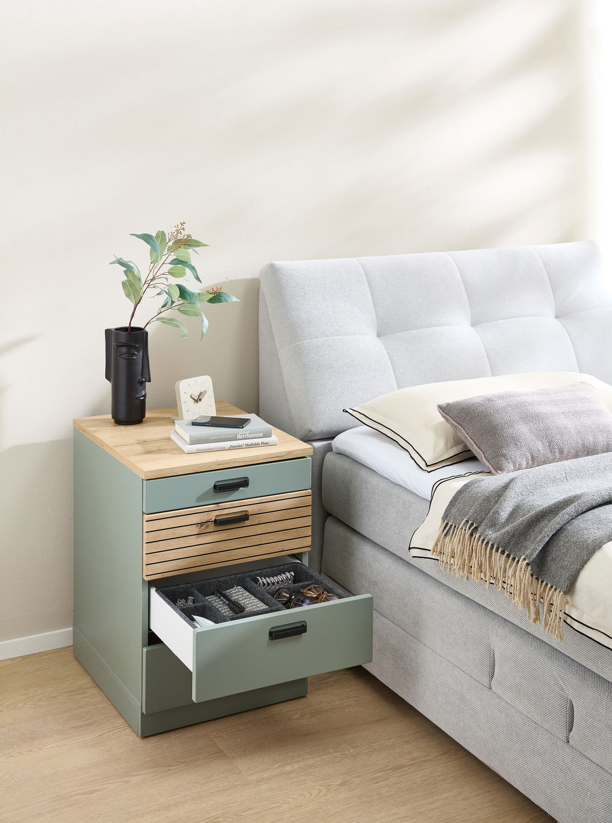 Rollcontainer mit drei Schubladen, aus Holz und Metall, neben einem Bett platziert. Perspektive von vorne.