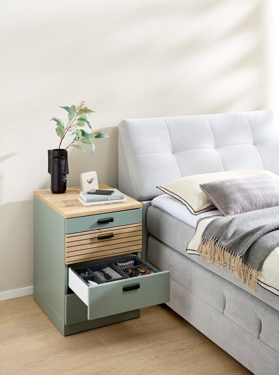 Rollcontainer mit drei Schubladen, aus Holz und Metall, neben einem Bett platziert. Perspektive von vorne.