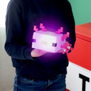 Een persoon houdt een lichtgevende, kubusvormige pixel-lamp in axolotl-stijl met roze kieuwen in zijn handen; vooraanzicht vanuit een licht schuin perspectief