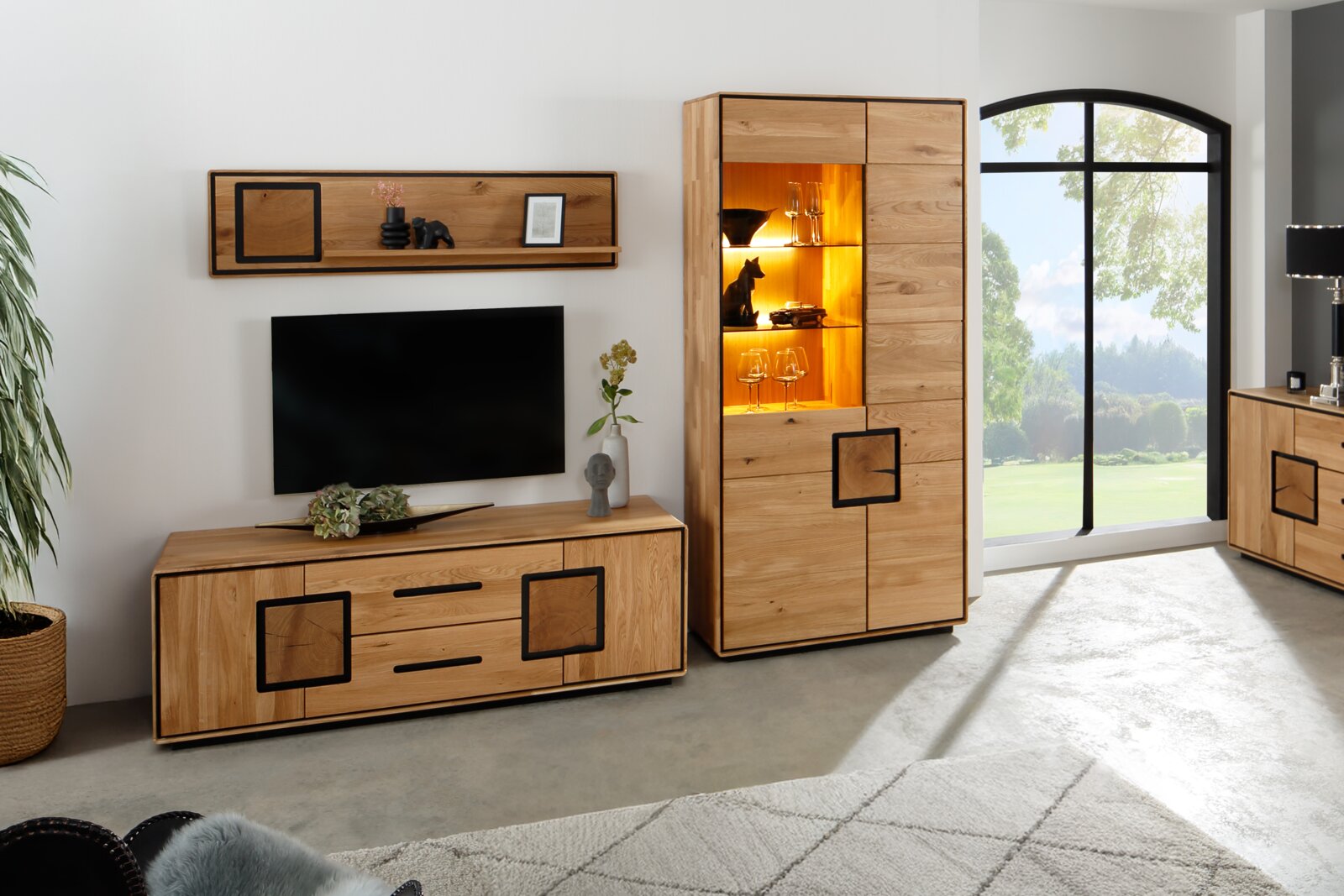 Wohnzimmermöbel aus massivem Holz: Lowboard mit Schubladen und Wandregal neben hoher Vitrine mit warm beleuchteten Glasböden, Frontansicht in leicht schräger Perspektive von links.