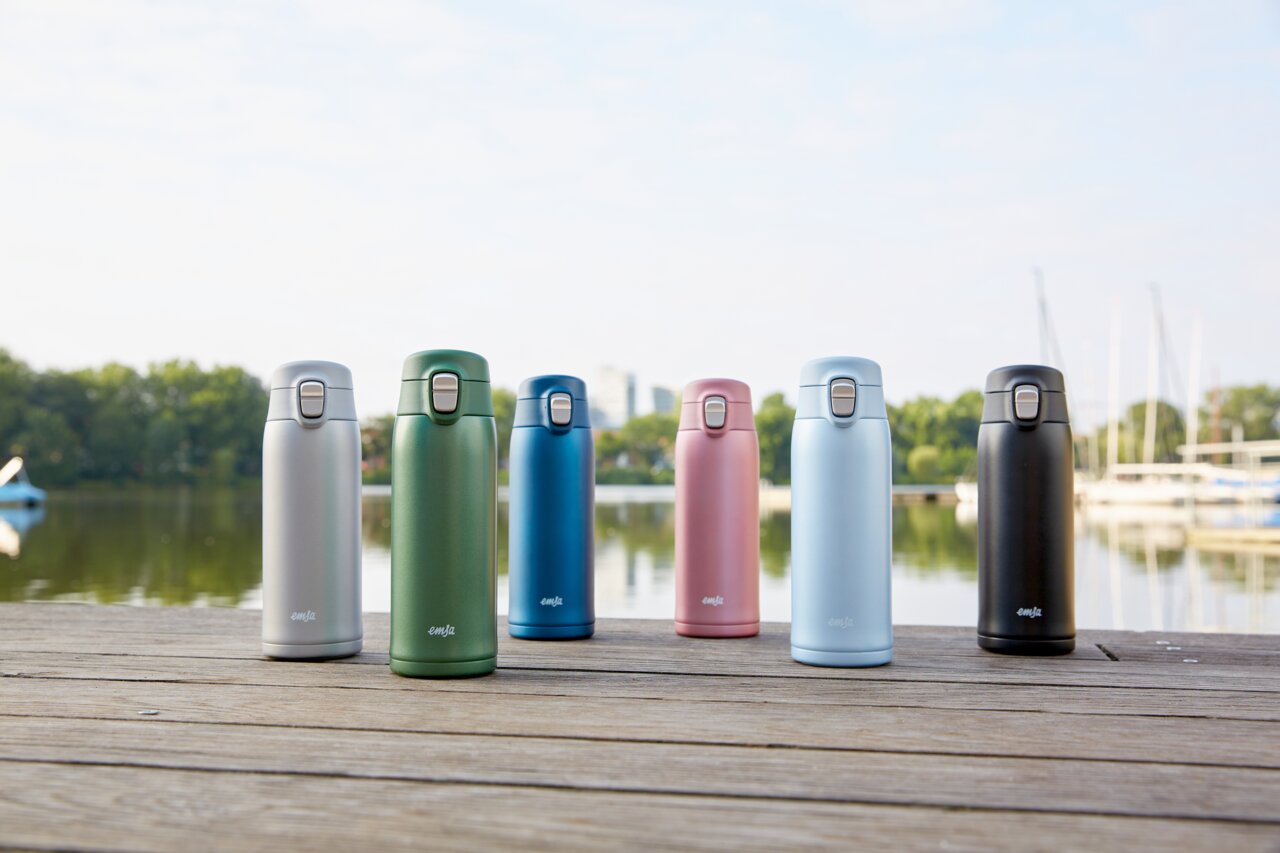 Sechs Thermobecher in verschiedenen Farben (Silber, Grün, Blau, Rosa, Hellblau, Schwarz) stehen auf einem Holzsteg mit einem See und Bäumen im Hintergrund, aus frontaler Perspektive fotografiert.