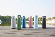 Sechs Thermobecher in verschiedenen Farben (Silber, Grün, Blau, Rosa, Hellblau, Schwarz) stehen auf einem Holzsteg mit einem See und Bäumen im Hintergrund, aus frontaler Perspektive fotografiert.