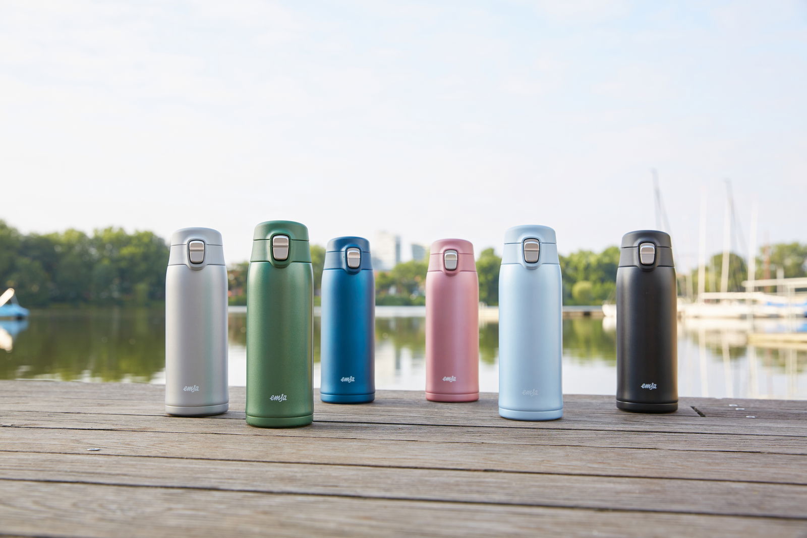 Sechs Thermobecher in verschiedenen Farben (Silber, Grün, Blau, Rosa, Hellblau, Schwarz) stehen auf einem Holzsteg mit einem See und Bäumen im Hintergrund, aus frontaler Perspektive fotografiert.