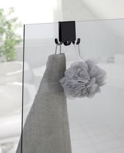 Doppel-Duschhaken aus Edelstahl, der an einer Glastür befestigt ist, mit einem Handtuch und einem Duschschwamm daran hängend. Perspektive von der Seite.