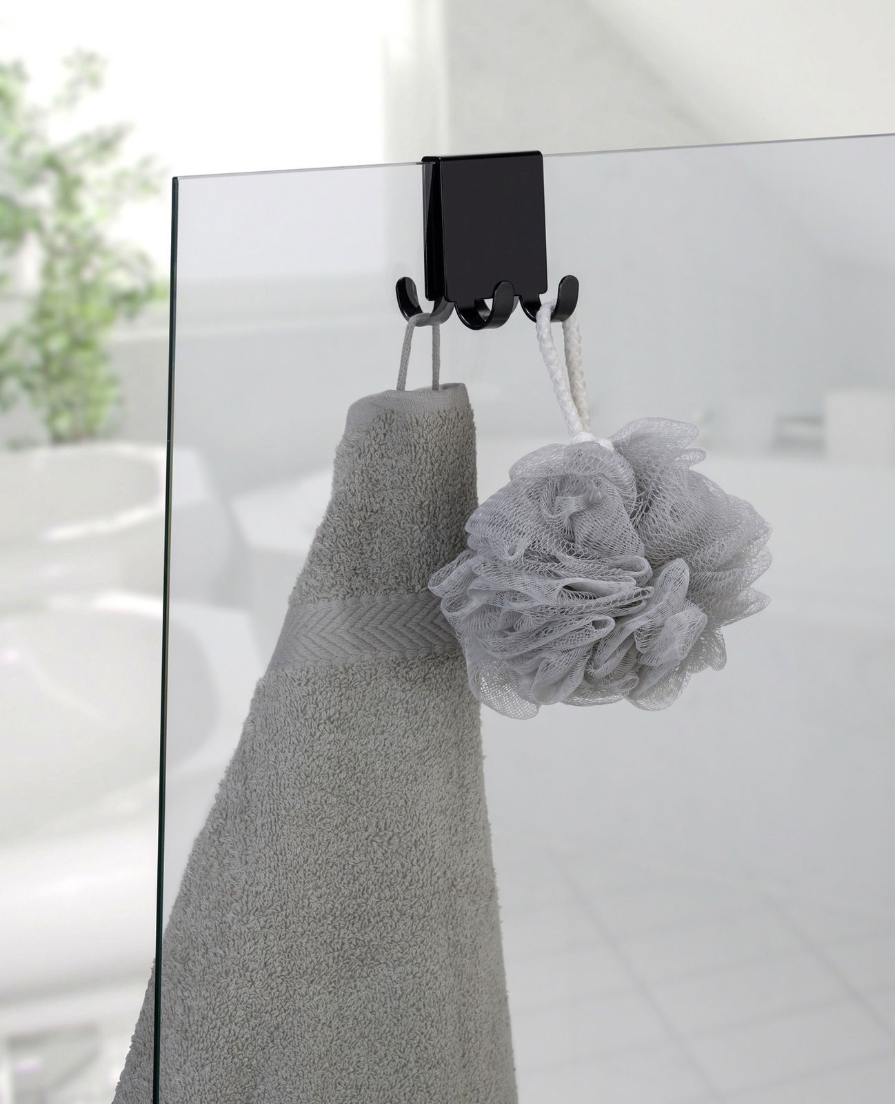 Doppel-Duschhaken aus Edelstahl, der an einer Glastür befestigt ist, mit einem Handtuch und einem Duschschwamm daran hängend. Perspektive von der Seite.