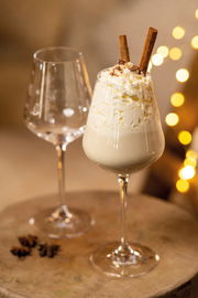 Zwei elegante Rotweinkelche, einer gefüllt mit einer cremigen weißen Getränkemischung, garniert mit Sahne und Zimtstangen, auf einem rustikalen Holztisch mit warmen Lichtern im Hintergrund. Seitliche Perspektive.