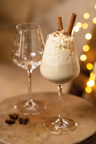 Zwei elegante Rotweinkelche, einer gefüllt mit einer cremigen weißen Getränkemischung, garniert mit Sahne und Zimtstangen, auf einem rustikalen Holztisch mit warmen Lichtern im Hintergrund. Seitliche Perspektive.