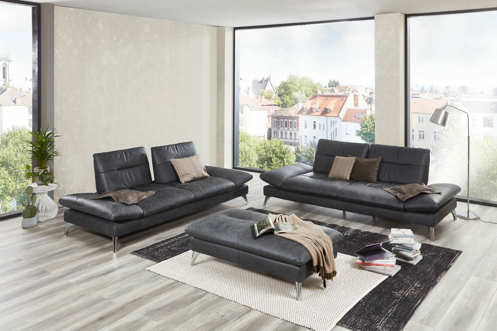 Modernes Wohnzimmer mit zwei dunkelgrauen 2,5-sitzigen Sofas und einem passenden Hocker, umgeben von großen Fenstern mit Stadtblick, aus der Perspektive von vorne.