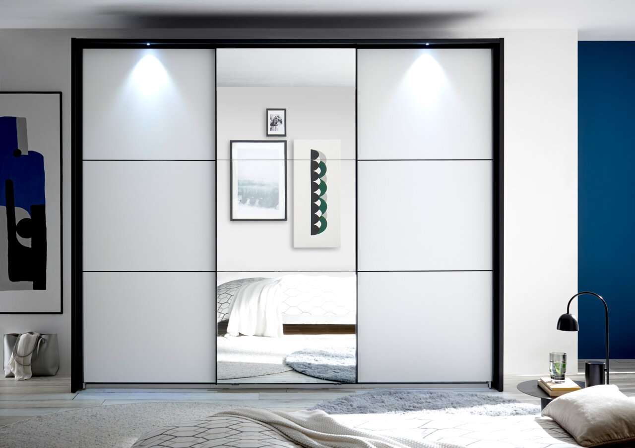 Moderne kledingkast met schuifdeuren en ingebouwde spiegel, gefotografeerd vanaf de voorkant. De kast staat in een stijlvolle ruimte met een blauwe muur en decoratieve elementen.