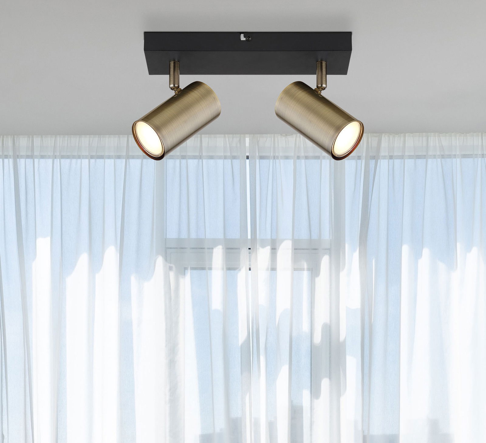 Globo Lighting Strahler 2-flg. ROBBY Zwei zylinderförmige Strahler in Altmessing-Optik, montiert an einer schwarzen Deckenhalterung, vor einem Fenster mit weißen Vorhängen. Perspektive von unten.