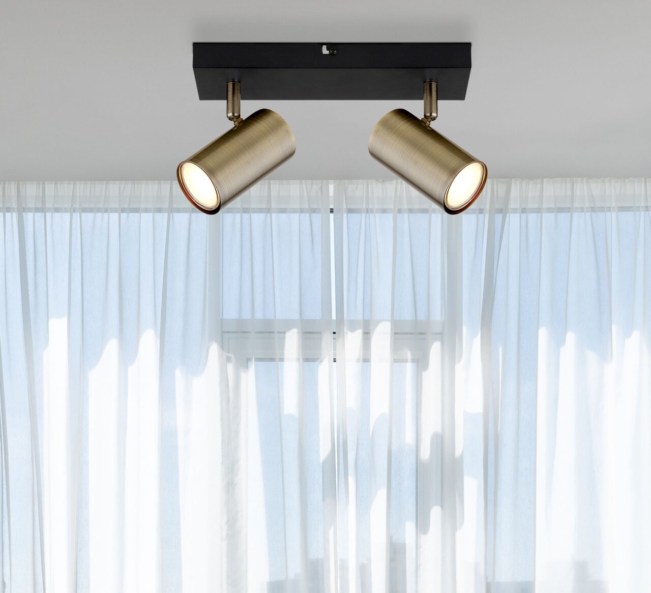 Globo Lighting Strahler 2-flg. ROBBY Zwei zylinderförmige Strahler in Altmessing-Optik, montiert an einer schwarzen Deckenhalterung, vor einem Fenster mit weißen Vorhängen. Perspektive von unten.