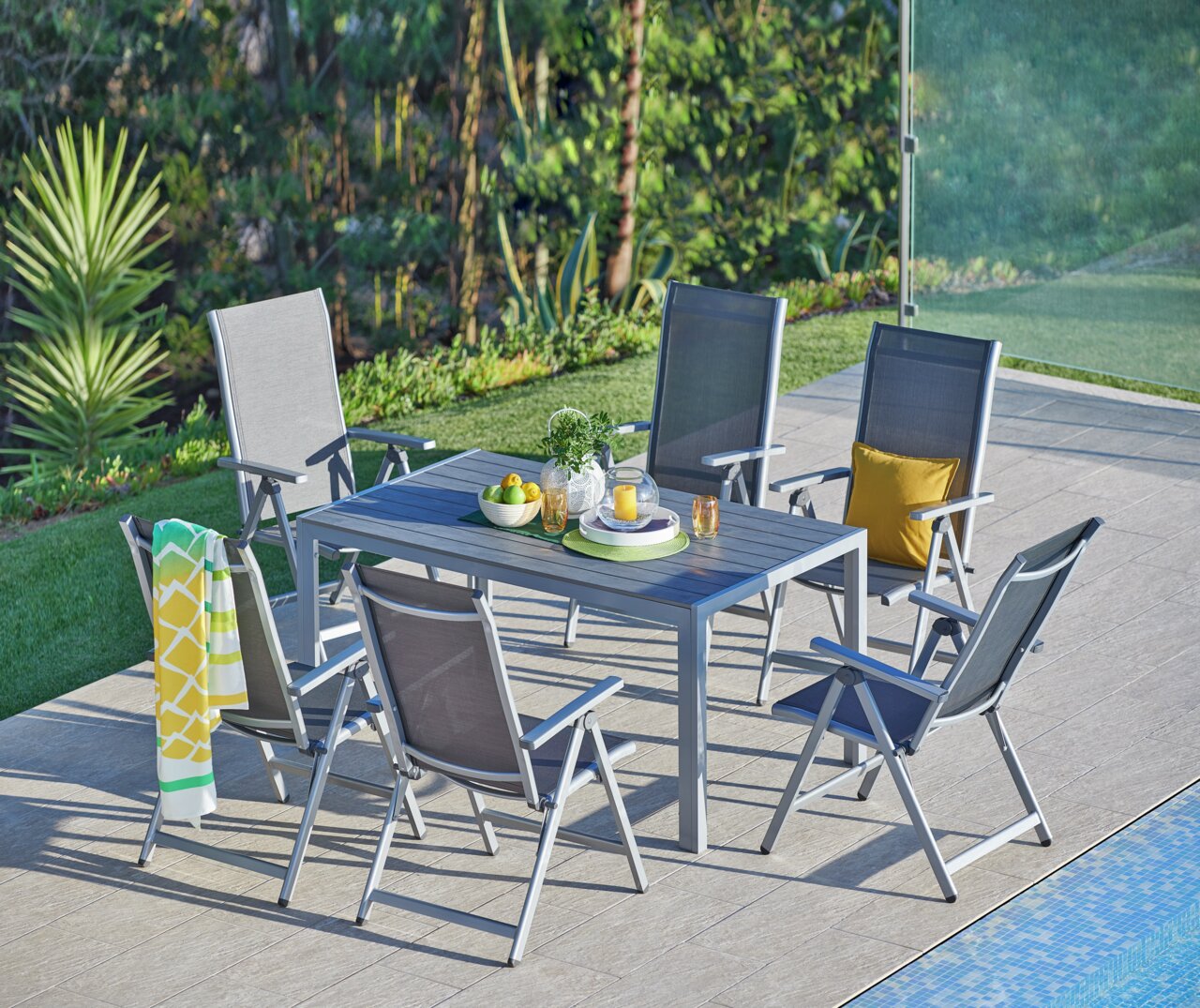 Gartenmöbel-Set mit sechs grauen Klappsesseln und einem Tisch auf einer Terrasse neben einem Pool, umgeben von grüner Vegetation, aus der Vogelperspektive.