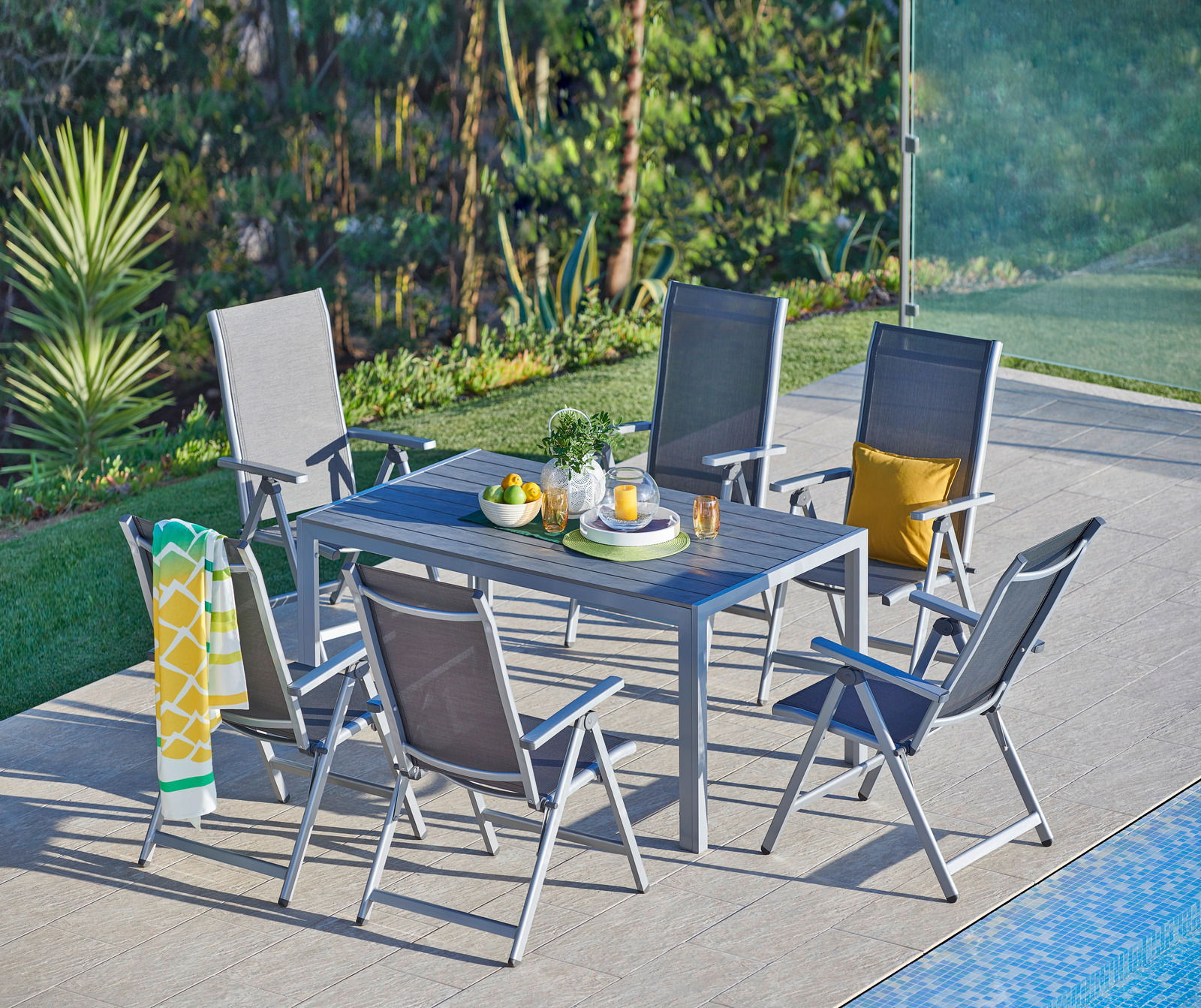 Gartenmöbel-Set mit sechs grauen Klappsesseln und einem Tisch auf einer Terrasse neben einem Pool, umgeben von grüner Vegetation, aus der Vogelperspektive.