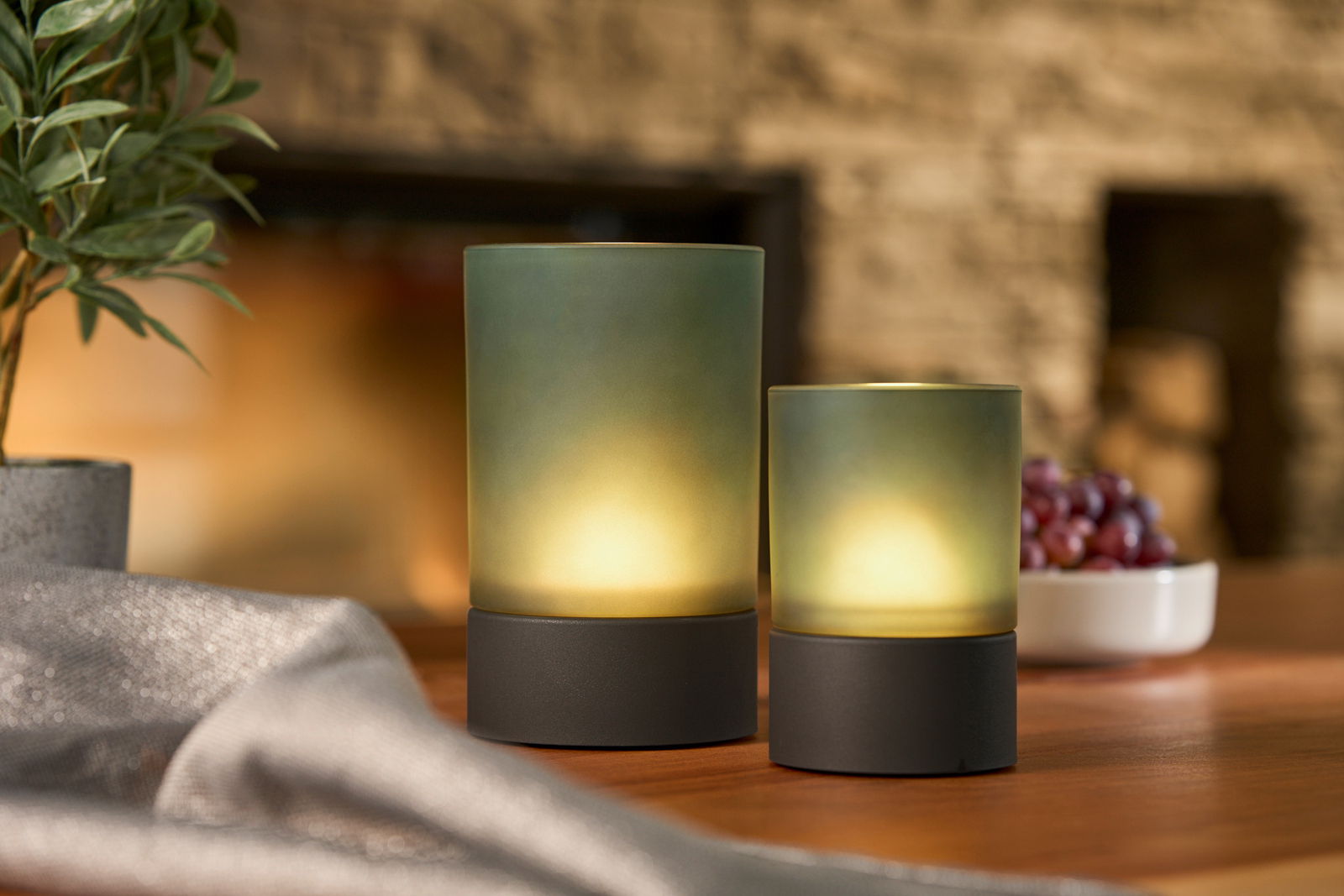 Zwei Akku-Windlichter in verschiedenen Größen auf einem Tisch, umgeben von einer Pflanze und einer Schale mit Trauben, aus seitlicher Perspektive fotografiert.