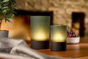 Zwei Akku-Windlichter in verschiedenen Größen auf einem Tisch, umgeben von einer Pflanze und einer Schale mit Trauben, aus seitlicher Perspektive fotografiert.