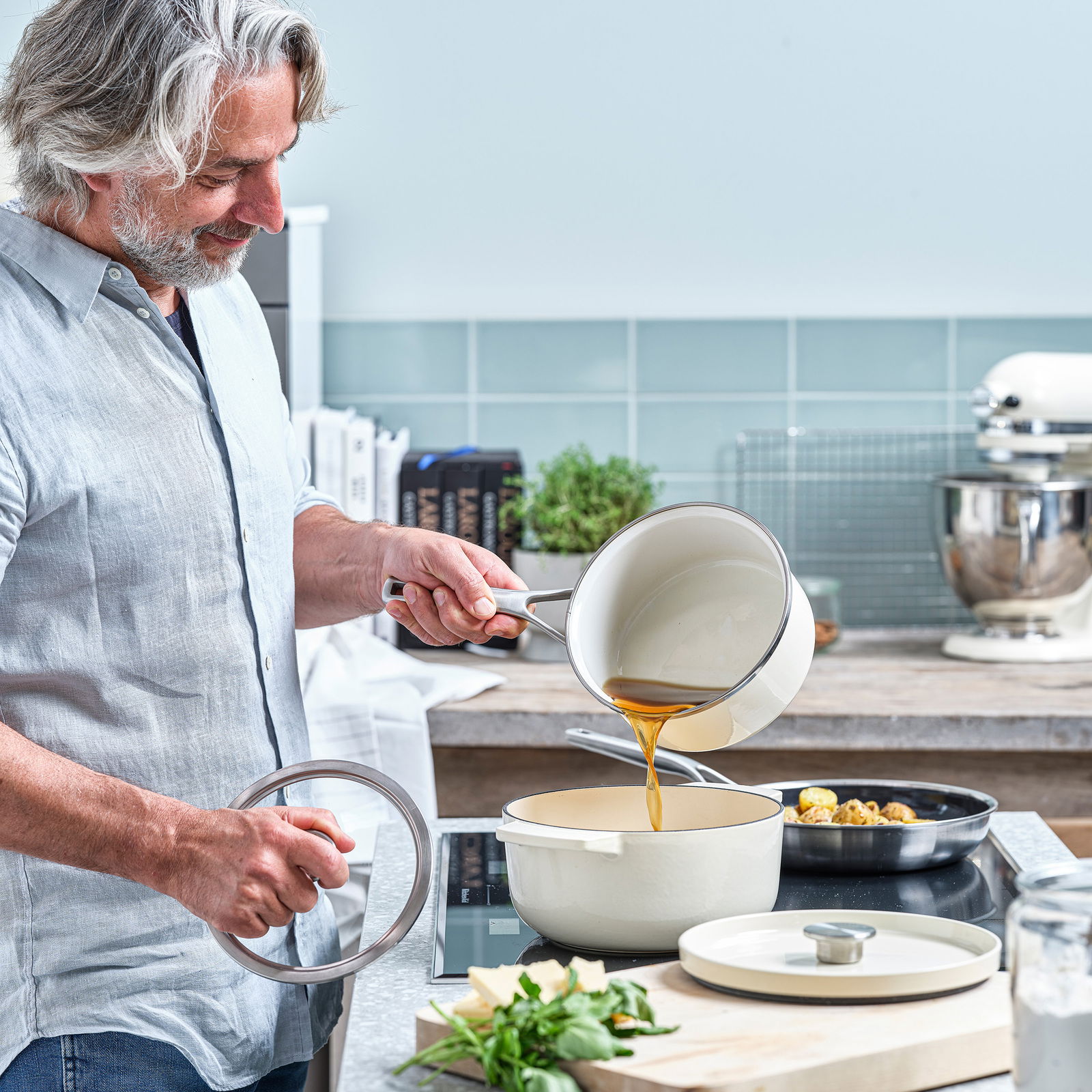 Ein Mann in einer modernen Küche gießt Flüssigkeit aus einem Topf in einen Gusseisen Bräter in der Farbe Mandel. Der Bräter steht auf einem Herd, und im Hintergrund sind Küchenutensilien und Kräuter zu sehen. Perspektive: seitlich.