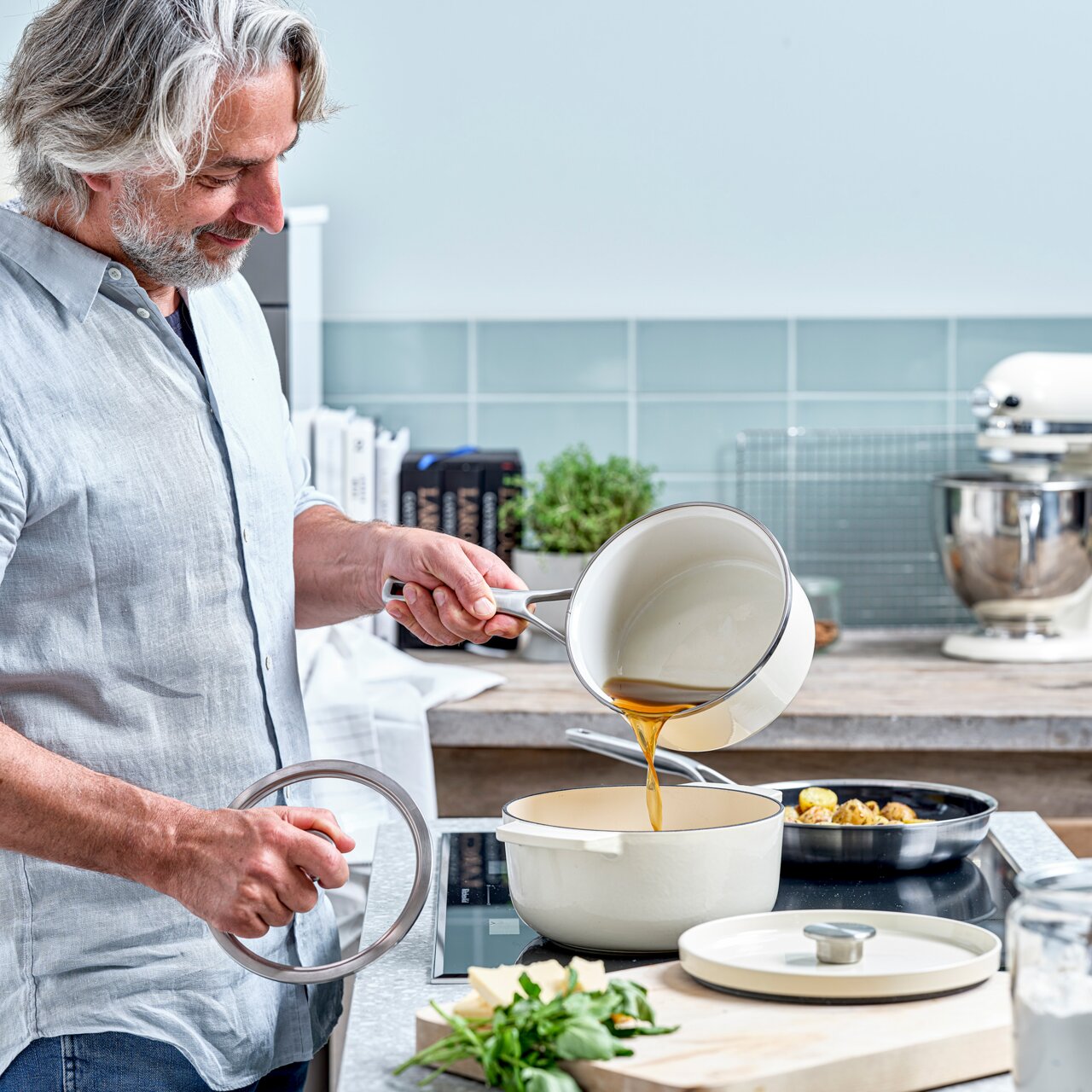 Ein Mann in einer modernen Küche gießt Flüssigkeit aus einem Topf in einen Gusseisen Bräter in der Farbe Mandel. Der Bräter steht auf einem Herd, und im Hintergrund sind Küchenutensilien und Kräuter zu sehen. Perspektive: seitlich.