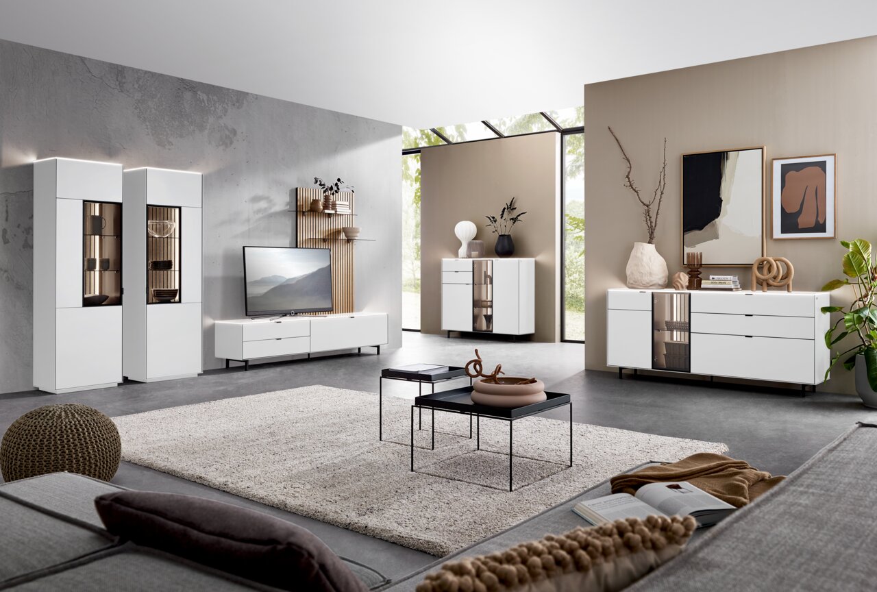 Moderne Wohnzimmermöbel in einem hellen Raum mit großen Fenstern, bestehend aus weißen Vitrinen, Sideboards und einem TV-Lowboard. Perspektive von der Sofaecke aus.