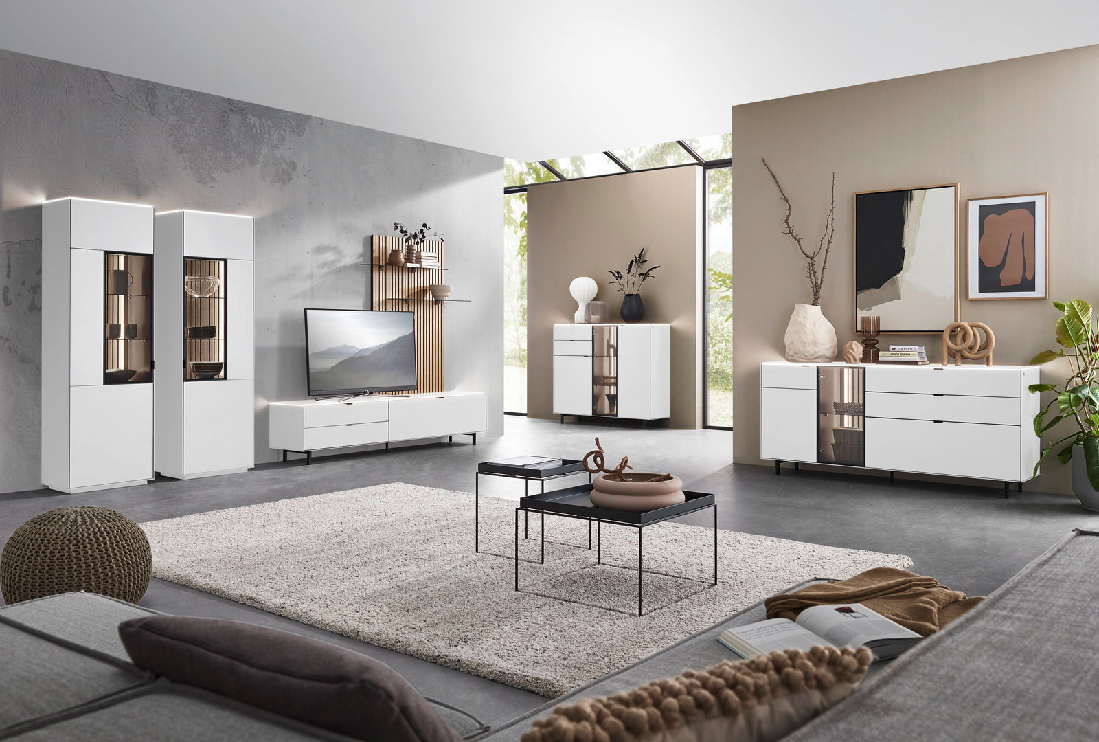 Moderne Wohnzimmermöbel in einem hellen Raum mit großen Fenstern, bestehend aus weißen Vitrinen, Sideboards und einem TV-Lowboard. Perspektive von der Sofaecke aus.