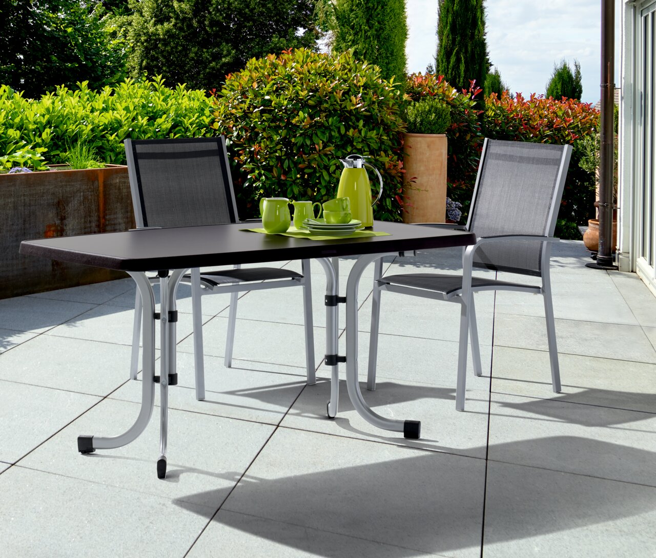 Klapptisch in Graphit auf einer Terrasse mit zwei Stühlen, von vorne betrachtet.