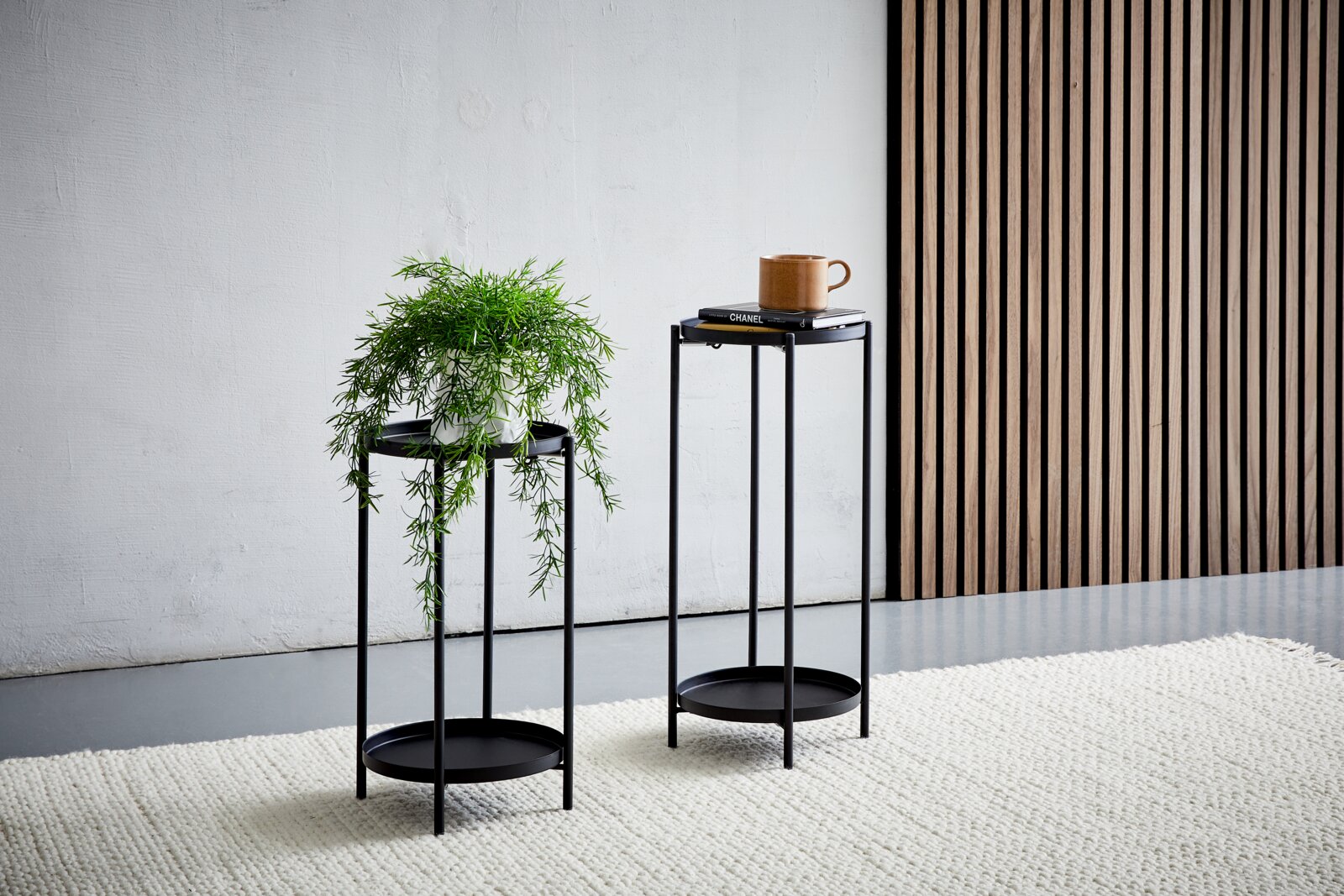Twee zwarte metalen bloemenpotten, de ene met een plant en de andere met een kopje en boeken, op een licht tapijt voor een wand met houten lambrisering, zijaanzicht.