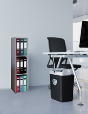 Moderner Aktenvernichter in Schwarz mit silbernem Kopf unter einem Büroschreibtisch, seitliche Perspektive im Büro neben einem Regal mit farbigen Ordnern