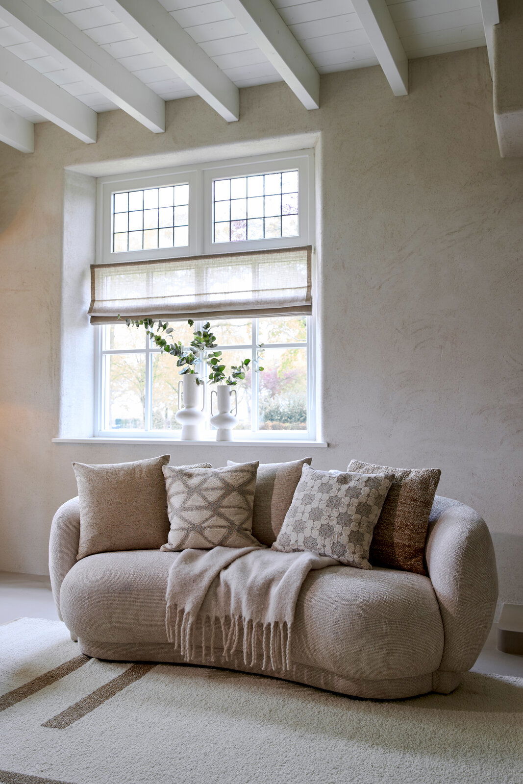 Gemütliches Wohnzimmer mit einem beigen Sofa, dekoriert mit vier stilvollen Kissen in neutralen Tönen, und einem weichen Überwurf. Im Hintergrund ein großes Fenster mit Pflanzen auf der Fensterbank. Perspektive: Frontalansicht auf das Sofa.