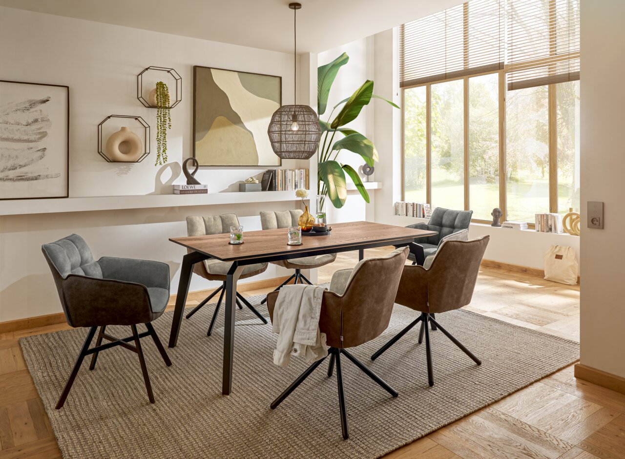 Moderner Esstisch aus Holz mit schwarzen Metallbeinen, umgeben von gepolsterten Stühlen in einem hellen, stilvoll eingerichteten Raum mit großen Fenstern und dekorativen Elementen. Perspektive von der Seite.