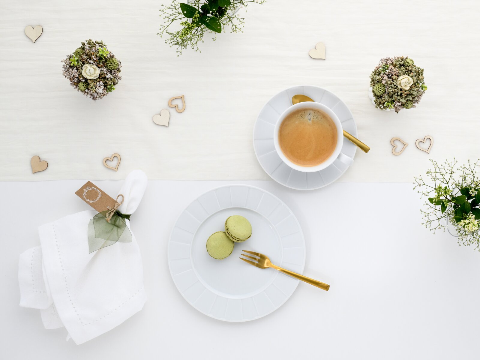 Draufsicht auf ein helles Tisch-Arrangement mit Espresso in weißer Tasse auf Untertasse mit goldenem Löffel, zwei grünen Macarons auf weißem Teller mit goldener Gabel, Serviette und Herz-Deko