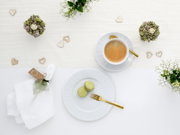 Draufsicht auf ein helles Tisch-Arrangement mit Espresso in weißer Tasse auf Untertasse mit goldenem Löffel, zwei grünen Macarons auf weißem Teller mit goldener Gabel, Serviette und Herz-Deko