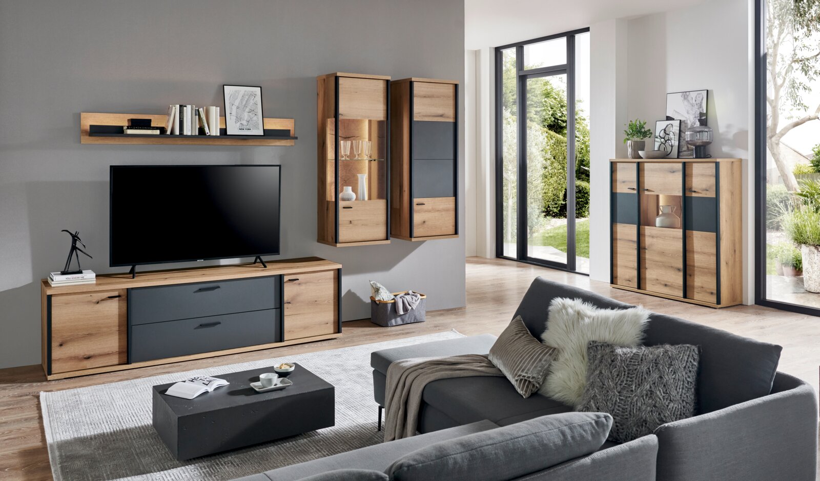 Modernes Wohnzimmer mit Wohnwand aus Holz und anthrazitfarbenen Fronten: Lowboard mit TV, Wandregal, zwei hängende Vitrinen und Highboard mit Beleuchtung; aufgenommen in schräger Frontalansicht aus dem Raum.