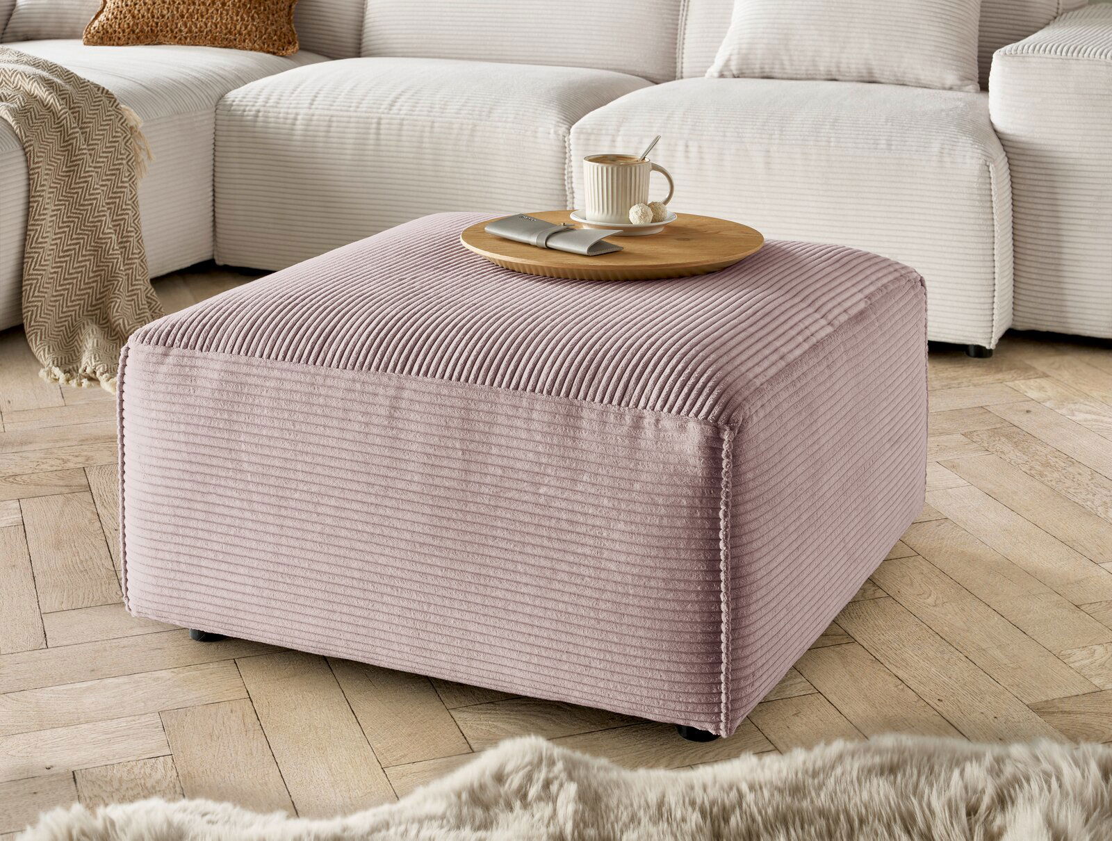 Hocker in rosa Cordstoff, quadratisch, mit einer Tasse und einem Teller darauf, in einem Wohnzimmer mit Holzboden, seitliche Perspektive