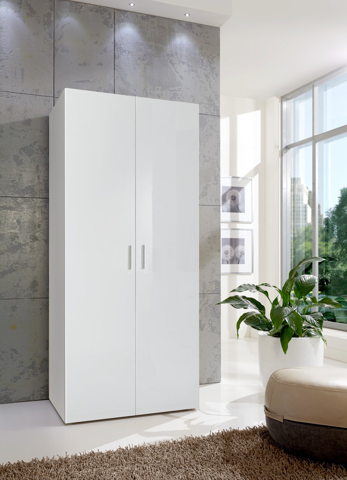 Moderner weißer Kleiderschrank mit zwei Türen, frontal fotografiert, in einem hellen Raum mit großen Fenstern und einer Pflanze im Vordergrund.