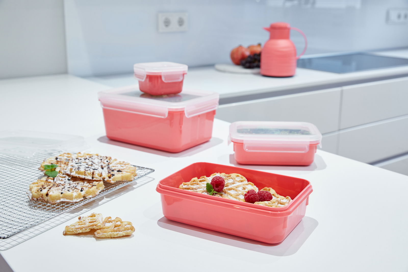Rote Frischhaltedosen in verschiedenen Größen auf einer Küchenarbeitsplatte, gefüllt mit Waffeln und Himbeeren, aus einer seitlichen Perspektive fotografiert.