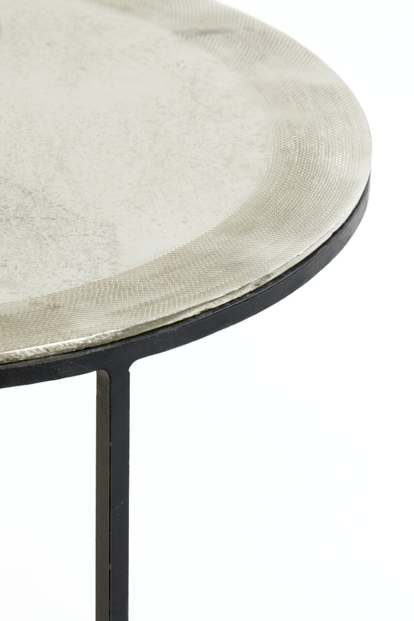 Close-up van een rond bijzettafeltje met een metalen oppervlak en een zwart onderstel, gezien vanaf de zijkant.