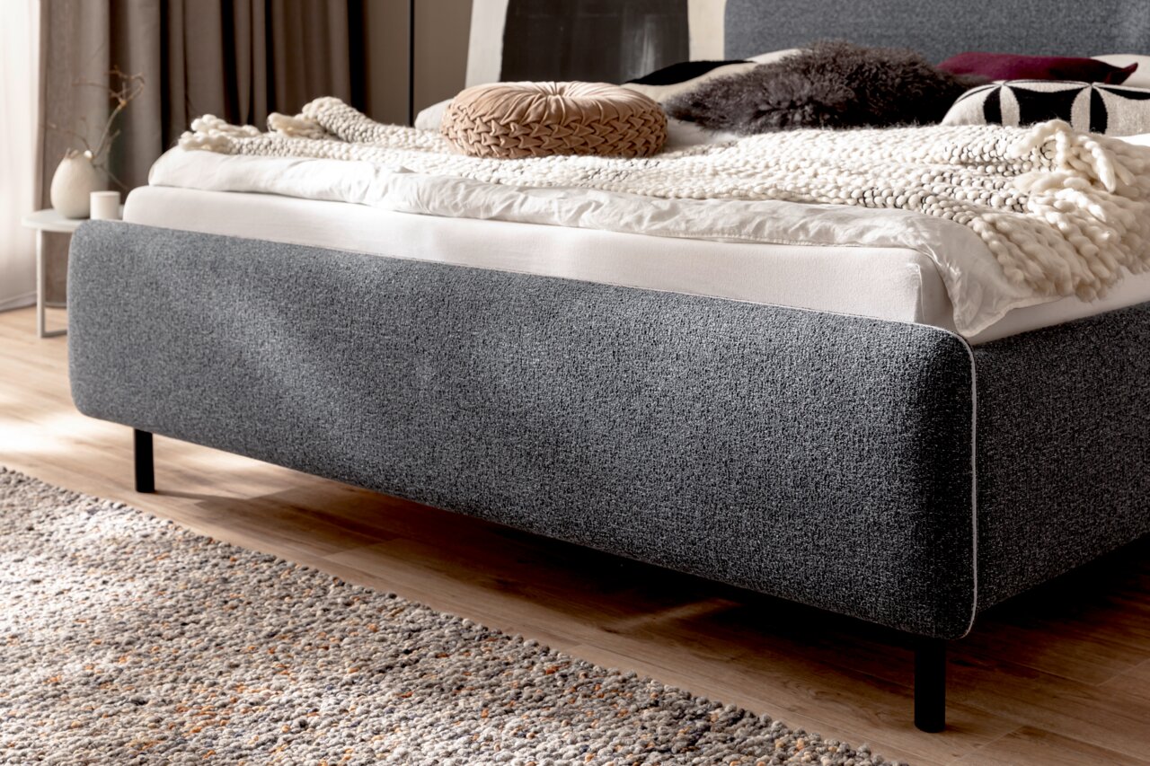 Seitliche Perspektive eines grauen Polsterbetts mit dekorativen Kissen und Decken auf einem Holzboden.