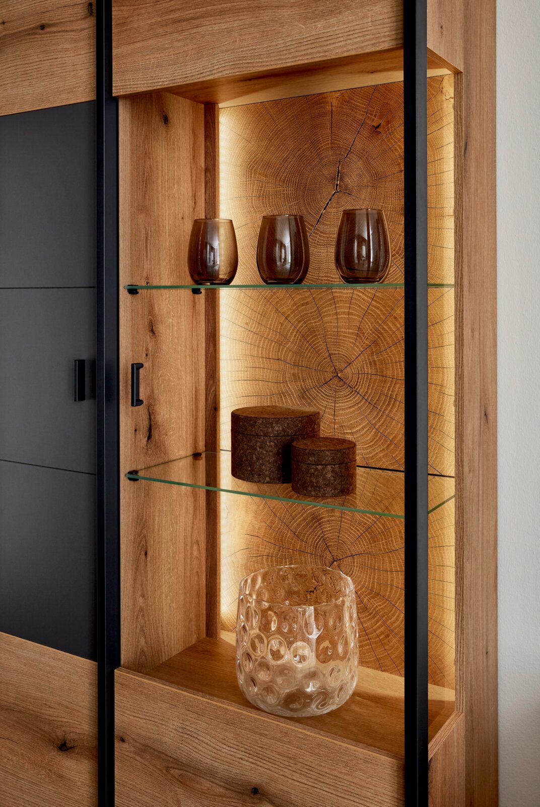 Detailansicht eines modernen Vitrinenschranks aus Holz mit schwarzem Rahmen, Glasböden und warm beleuchteter Rückwand in Baumstamm-Optik; frontale Perspektive mit leichtem Blick von links, dekoriert mit Gläsern und Dosen.