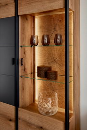 Detailansicht eines modernen Vitrinenschranks aus Holz mit schwarzem Rahmen, Glasböden und warm beleuchteter Rückwand in Baumstamm-Optik; frontale Perspektive mit leichtem Blick von links, dekoriert mit Gläsern und Dosen.