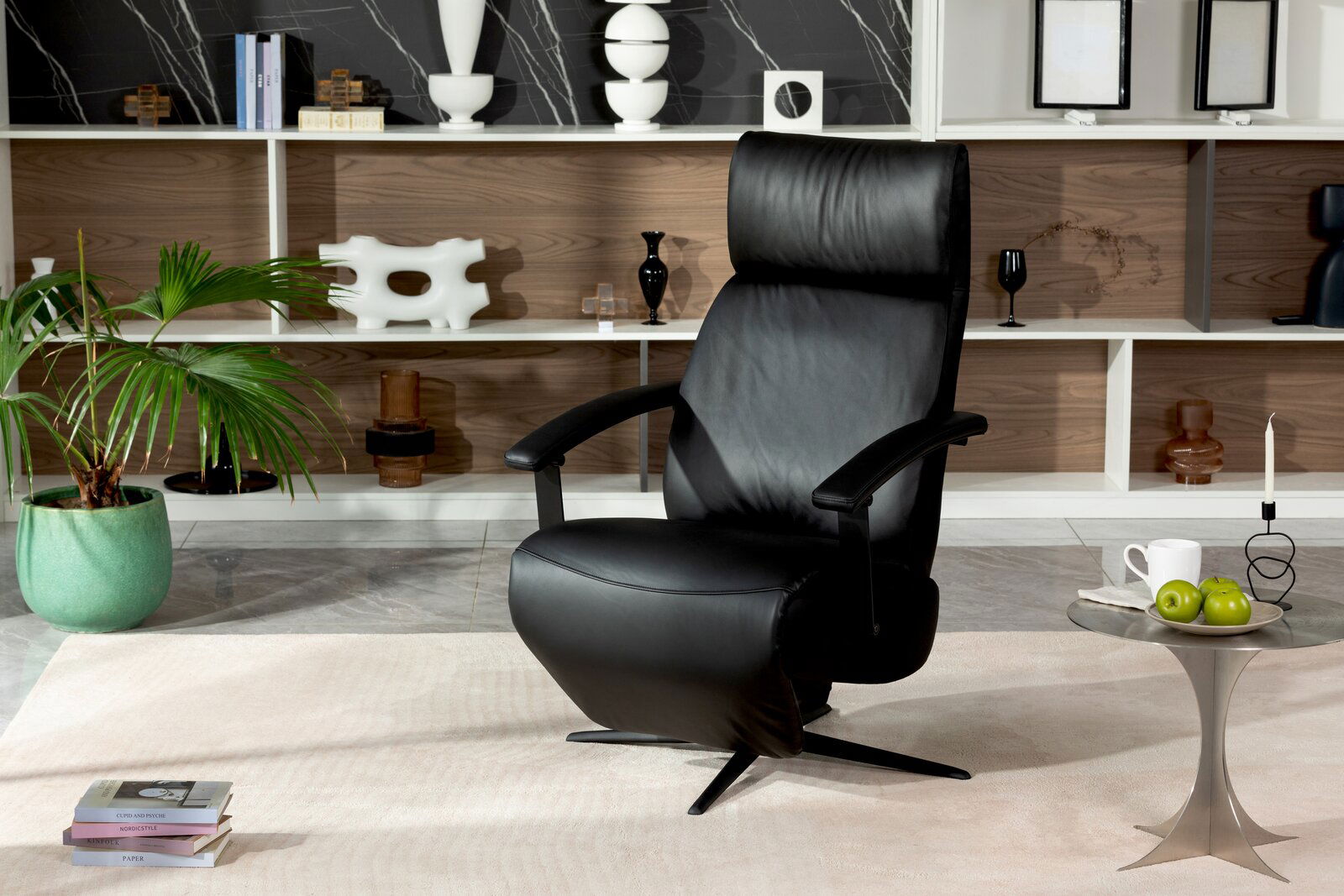 Schwarzer Relaxsessel aus Leder in einem modernen Wohnzimmer, frontal fotografiert. Im Hintergrund ein Regal mit Dekorationen und eine grüne Pflanze.