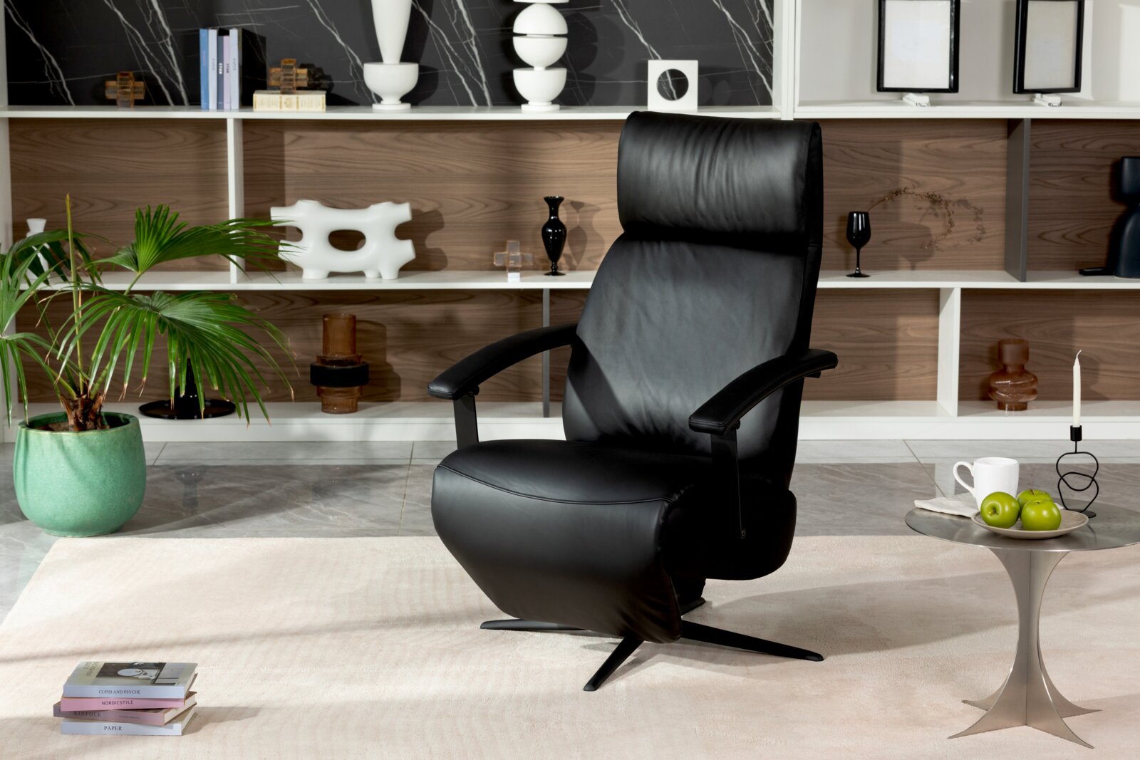XORA Relaxsessel MAX 2.0 Schwarzer Relaxsessel aus Leder in einem modernen Wohnzimmer, frontal fotografiert. Im Hintergrund ein Regal mit Dekorationen und eine grüne Pflanze.