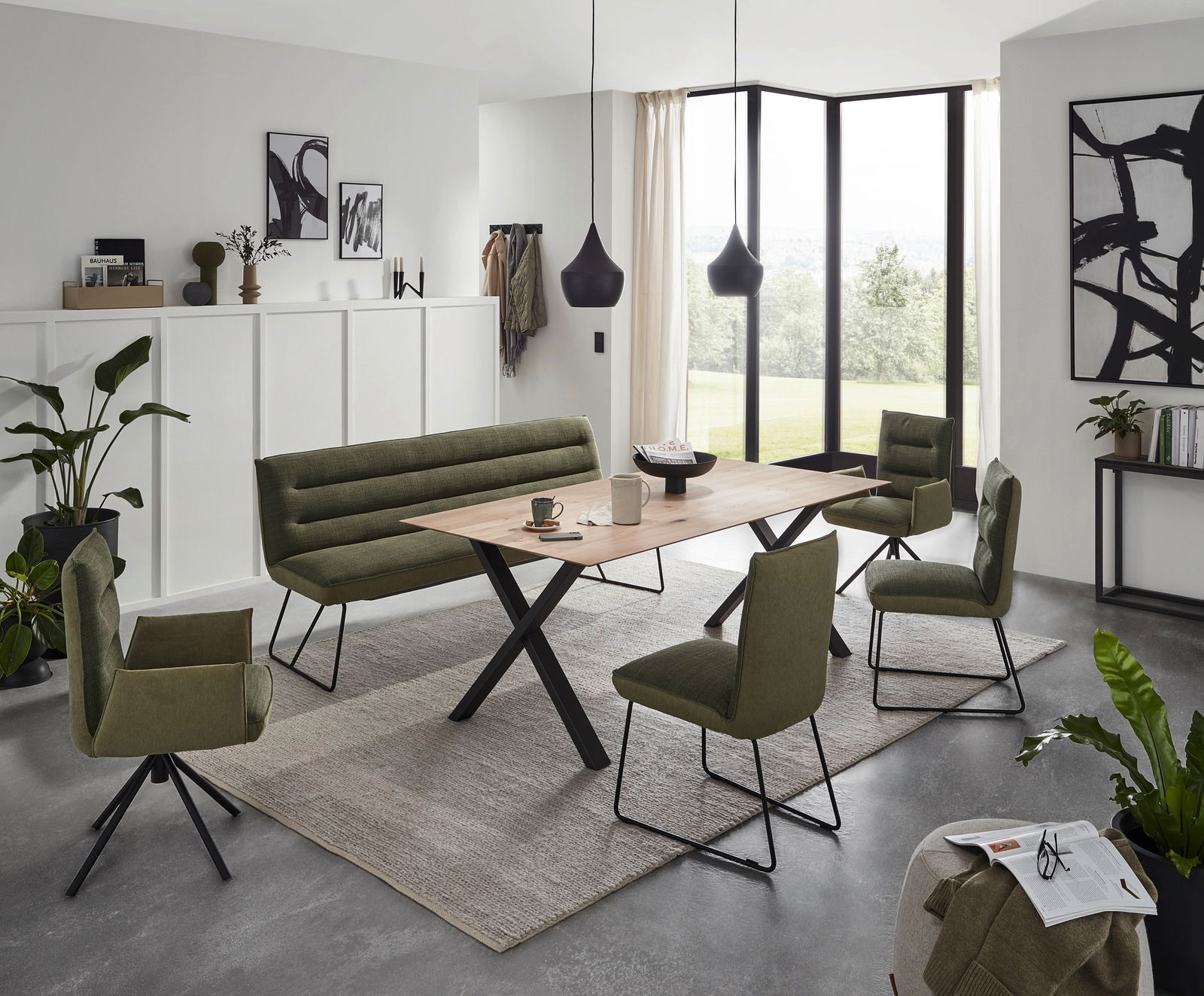 Dieter Knoll Bank ABANO Moderne Esszimmermöbel in einem hellen Raum mit großen Fenstern, bestehend aus einem Holztisch mit X-förmigen Beinen, umgeben von grünen Polsterstühlen und einer passenden Bank, aus der Perspektive des Raumes betrachtet.