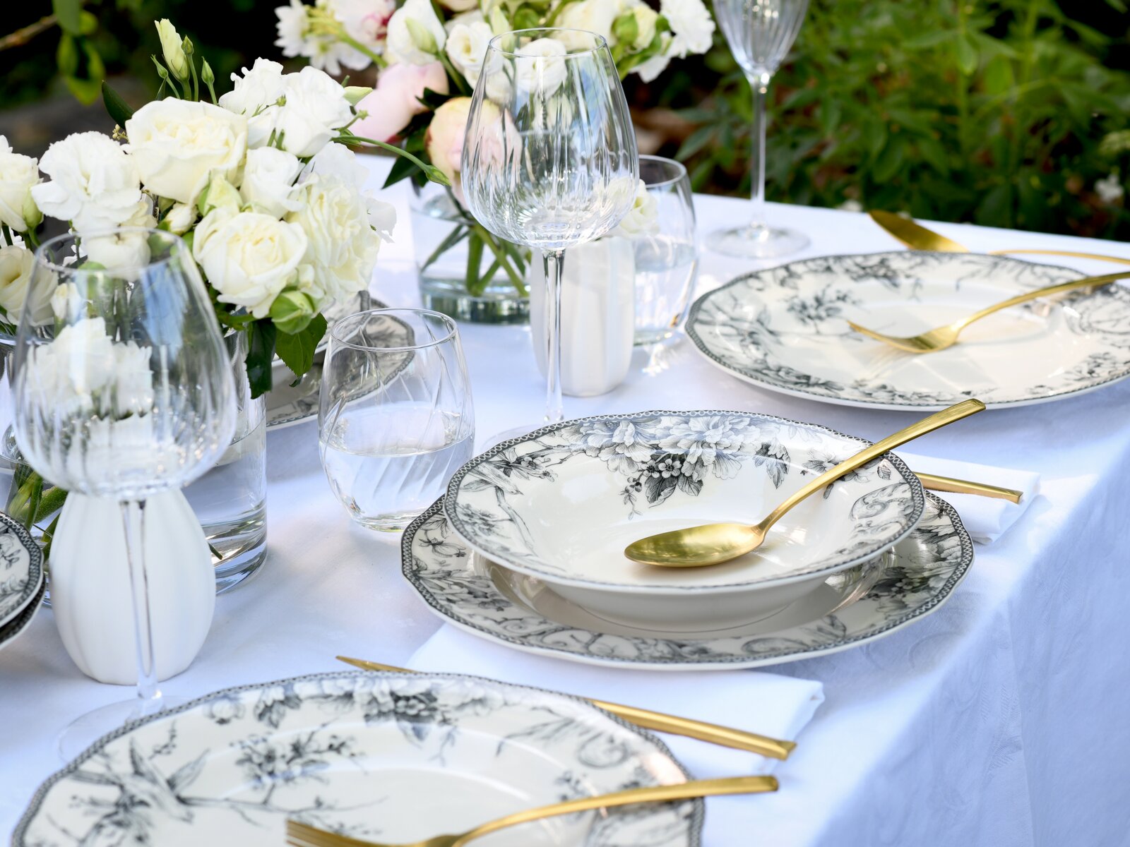 Elegant gedeckter Outdoor-Tisch mit weißer Tischdecke, schwarz-weiß floral gemustertem Porzellan, goldfarbenem Besteck, klaren Wein- und Wassergläsern sowie weißen Blumenarrangements; Perspektive: leicht schräg von oben in Nahaufnahme.