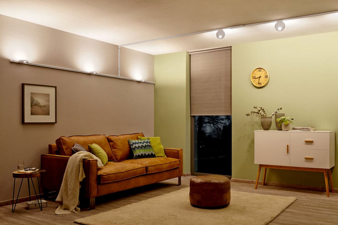 Wohnzimmer mit moderner Beleuchtung, die an der Decke entlang verläuft, mit einem Flexverbinder für URAIL-System. Perspektive von der Seite des Raumes mit Sofa, Beistelltisch und Kommode.