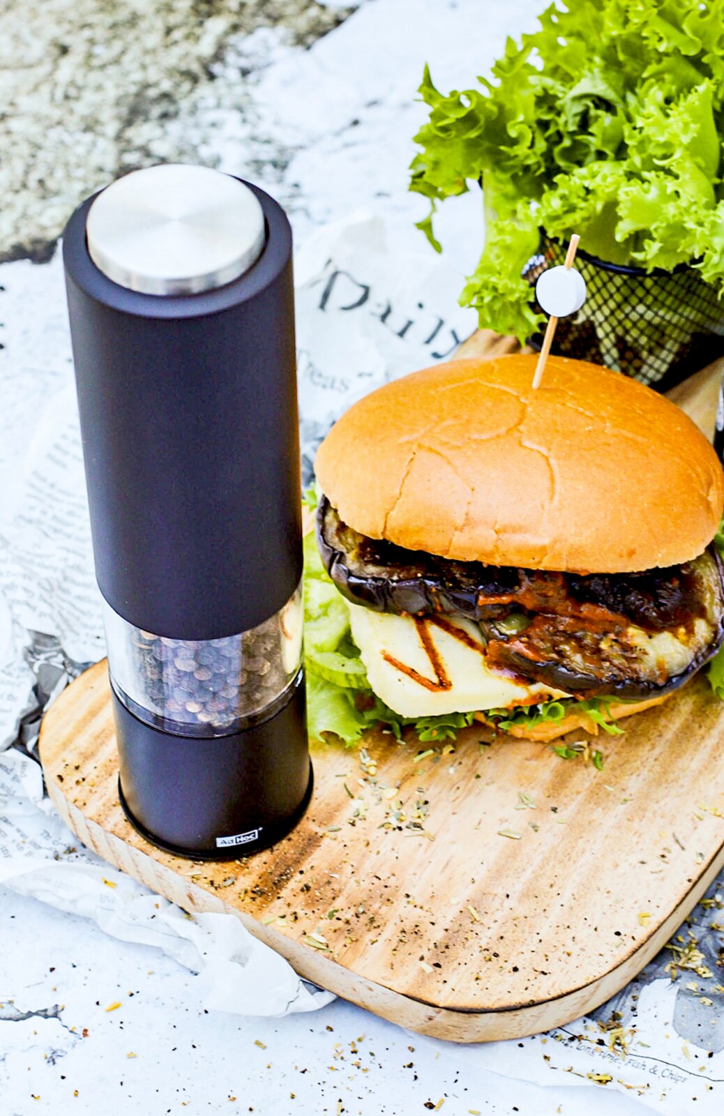 Elektrische Pfeffer- oder Salzmühle in schwarzem Design, stehend auf einem Holzbrett neben einem Burger, aus seitlicher Perspektive fotografiert.