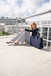 Frau sitzt auf einer Terrasse mit einer großen, dunkelblauen, gesteppten Tasche neben sich. Die Tasche hat zwei Henkel und steht aufrecht auf dem Boden.