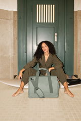 Frau sitzt vor einer grünen Tür mit einer großen, grünen Tasche im Vordergrund. Perspektive von vorne.