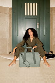 Frau sitzt vor einer grünen Tür mit einer großen, grünen Tasche im Vordergrund. Perspektive von vorne.